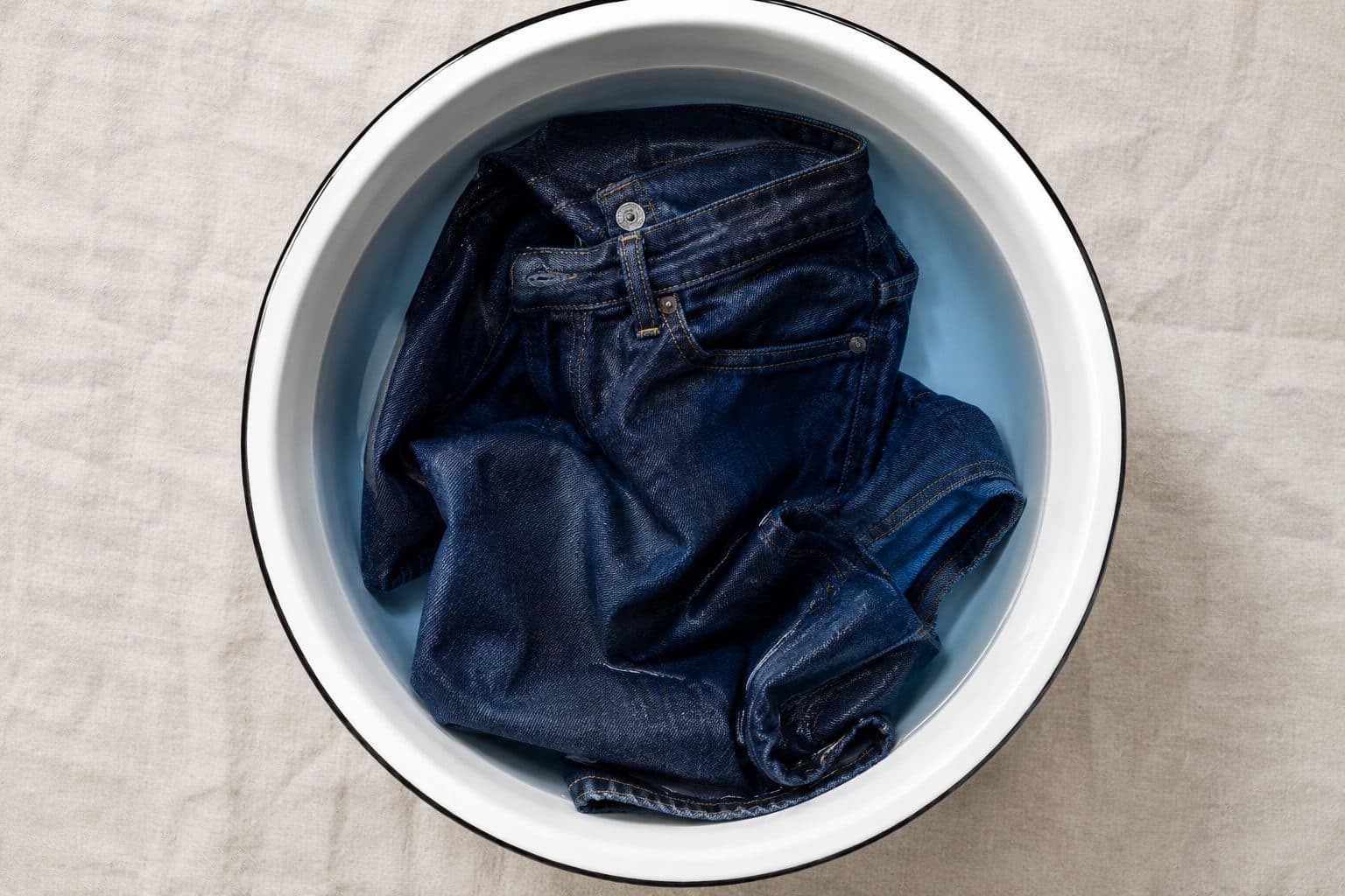 A single pair of dark indigo trousers alone in a white enamel basin of faintly blue water
