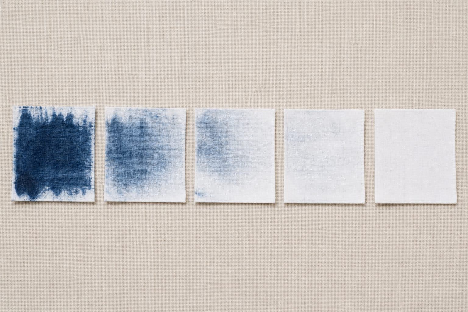 Five white cotton test cloths in a row, the left one heavily stained with blue indigo and each one to the right progressively cleaner until the last is white