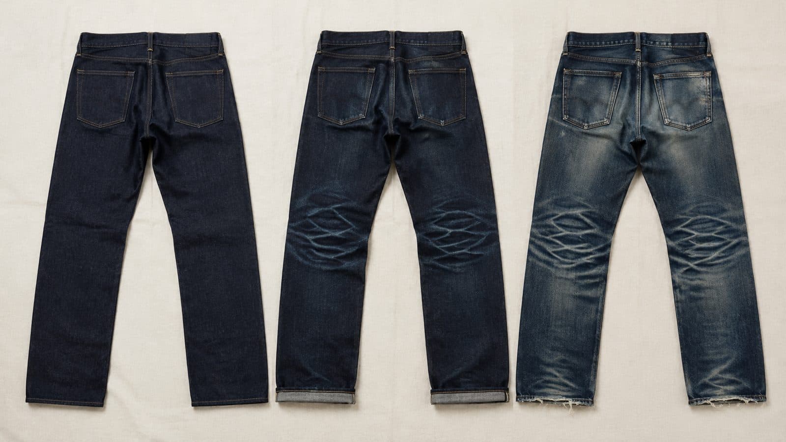 Three identical pairs of unwashed denim trousers side by side: one brand new and uniformly dark, one with faint creases behind the knee, one heavily faded with a worn seat and frayed hems