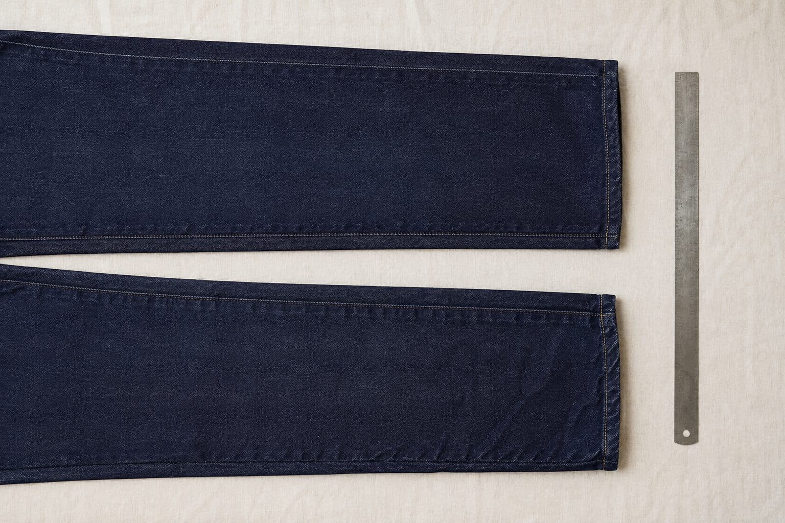 Two identical denim trouser legs laid parallel with a tailor's ruler, the lower one visibly shorter after washing